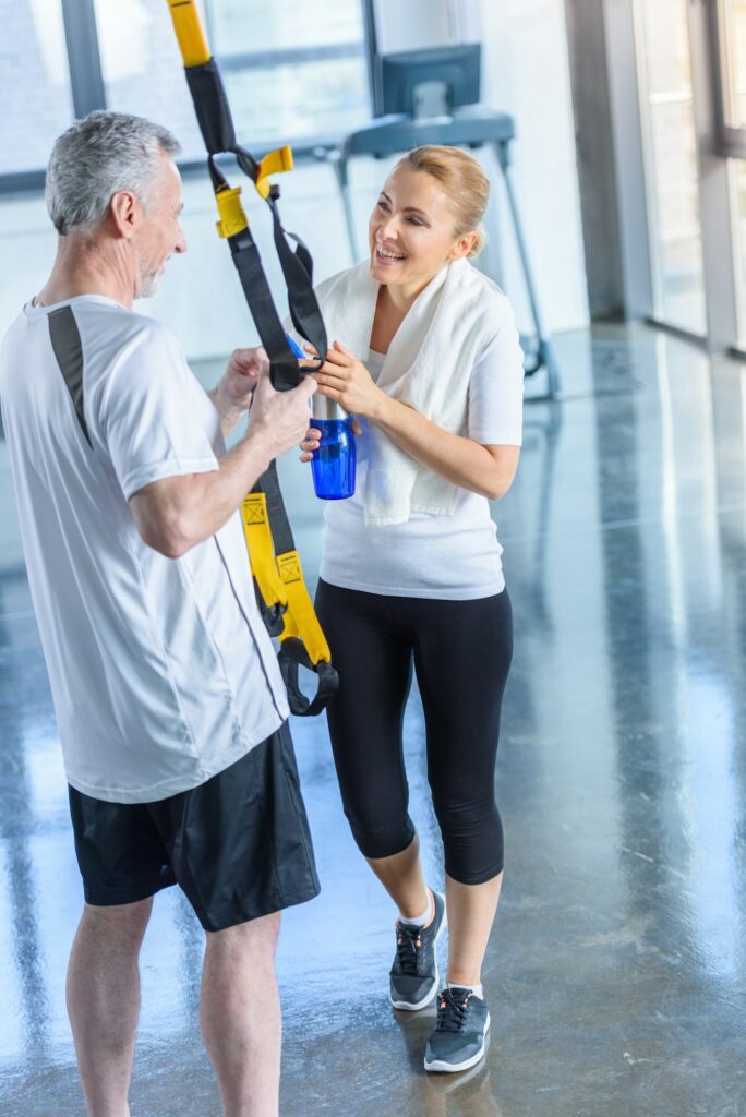 sportswoman holding sport bottle and senior sportsman with resistance band talking in sports center